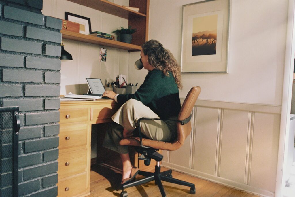 Empreendedor visto de costas em home office, junto a uma grande janela com vista da cidade, analisando documentos e conta bancária de campanha no laptop.