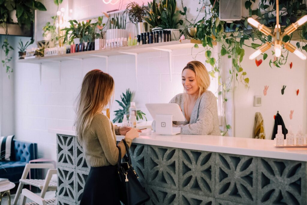 Interior de salão de beleza ou barbearia em tons pastel, com a proprietária organizando a recepção e materiais administrativos antes de contratar um funcionário.