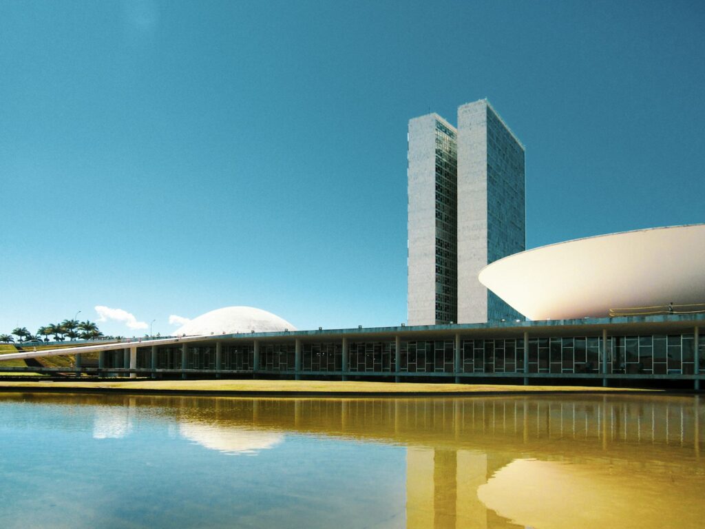 Exterior de prédio corporativo moderno em Brasília, com céu azul e arquitetura geométrica, enquanto profissionais analisam documentos sobre a transição da Reforma Tributária.