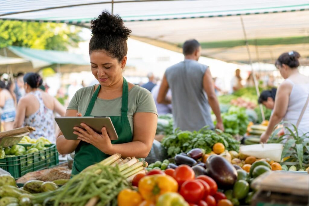 Vendedor em feira brasileira usando tablet em banca com frutas e verduras coloridas.